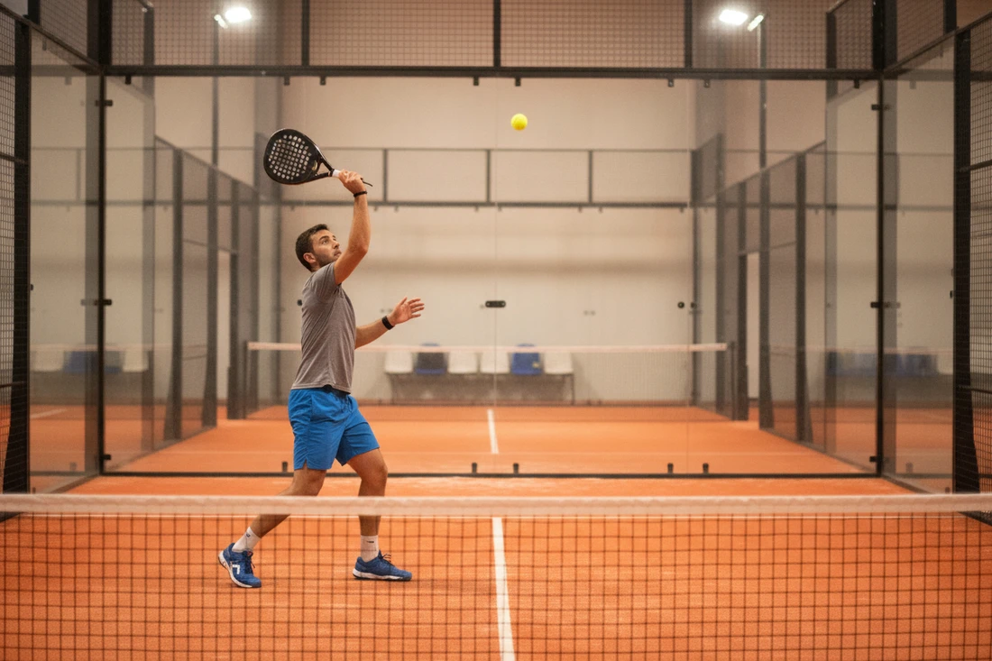 Joueur de padel côté gauche attaquant une balle haute au centre, terrain indoor orange