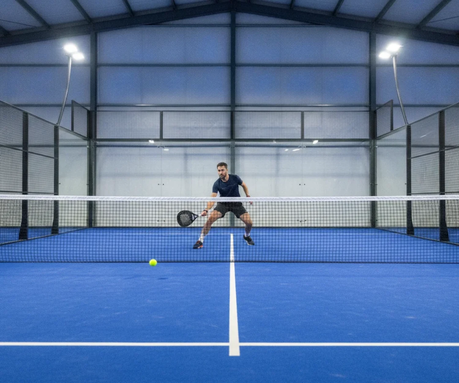 Joueur de padel jouant une balle basse vers les pieds adverses, terrain indoor bleu