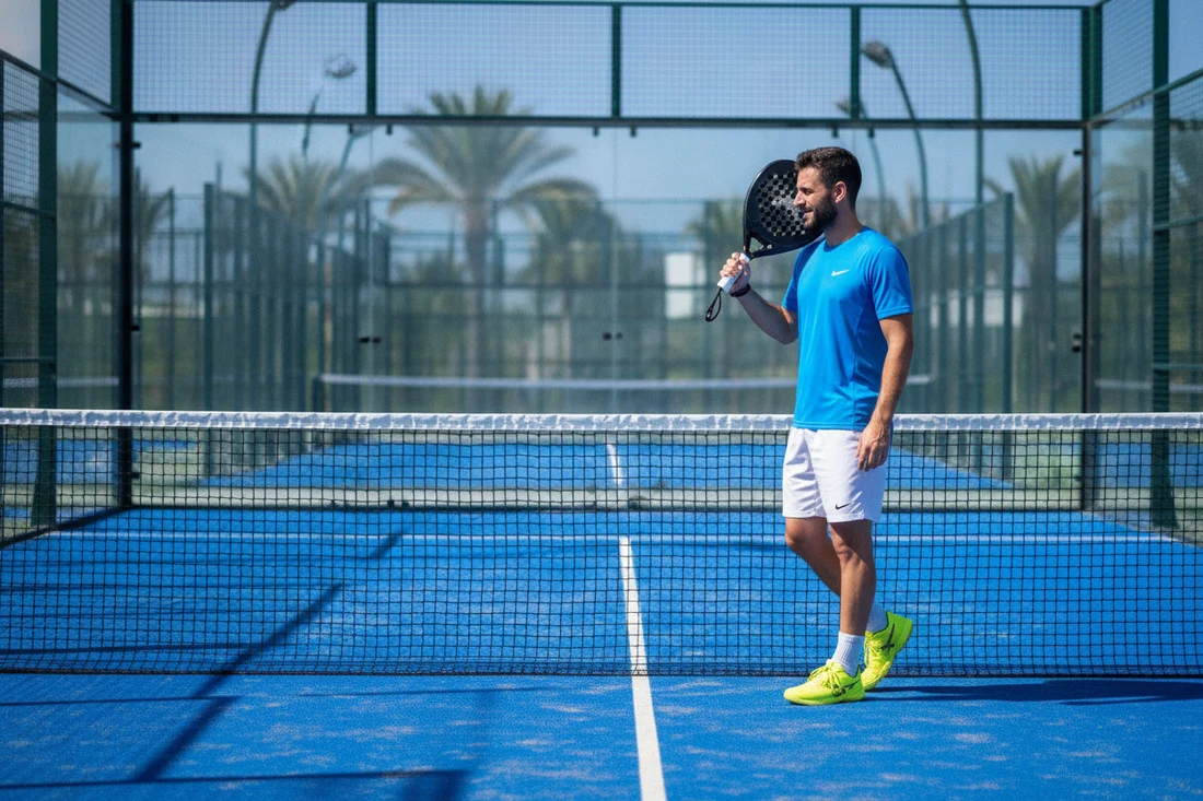 Joueur de padel confiant debout, raquette posée sur l'épaule au bord du filet sur court bleu