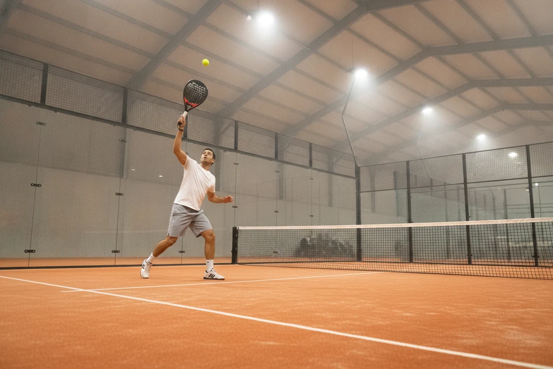 Joueur de padel jouant un lob sur une piste couverte au sol orange, clubs de padel à Brest