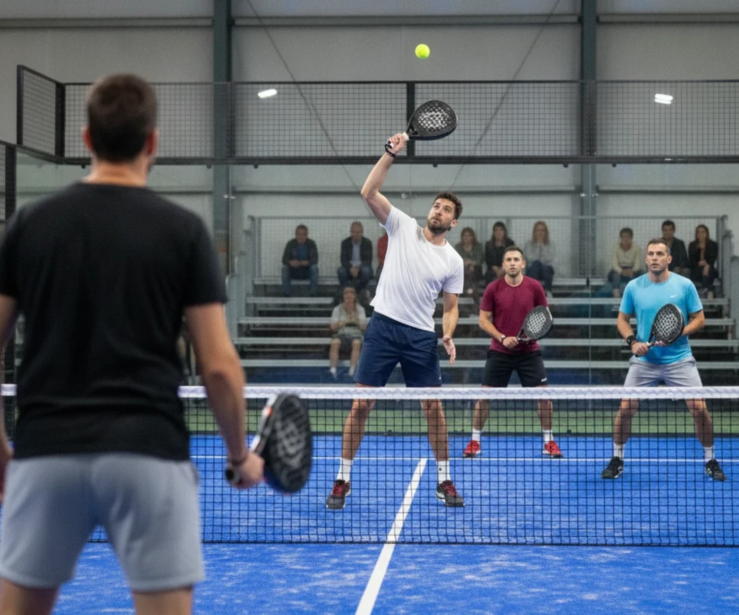 Un joueur de padel frappe une volée haute au filet devant une tribune de spectateurs dans une salle, partenaire flou au premier plan, piste bleue