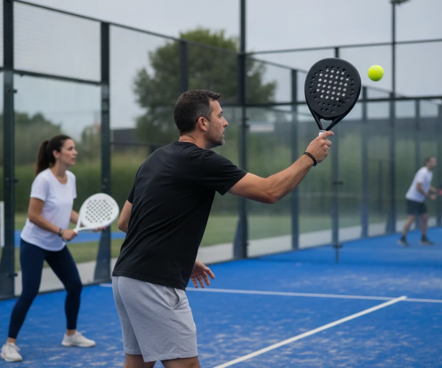 Un joueur de padel frappe une bandeja de profil sur un court extérieur bleu, sa partenaire en attente derrière lui