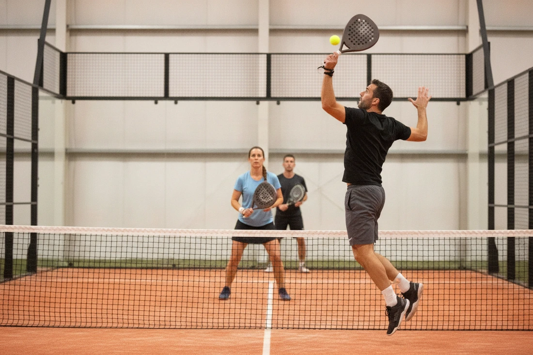 Un joueur de padel s'élève pour un smash au filet sur une piste orange, deux adversaires en position de défense