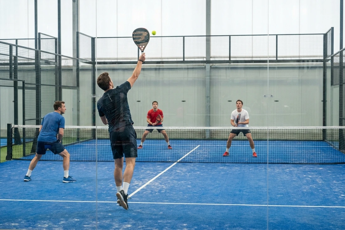 Joueur de padel au smash face à ses adversaires pendant une partie en double sur une piste couverte au sol bleu, clubs de la métropole nantaise