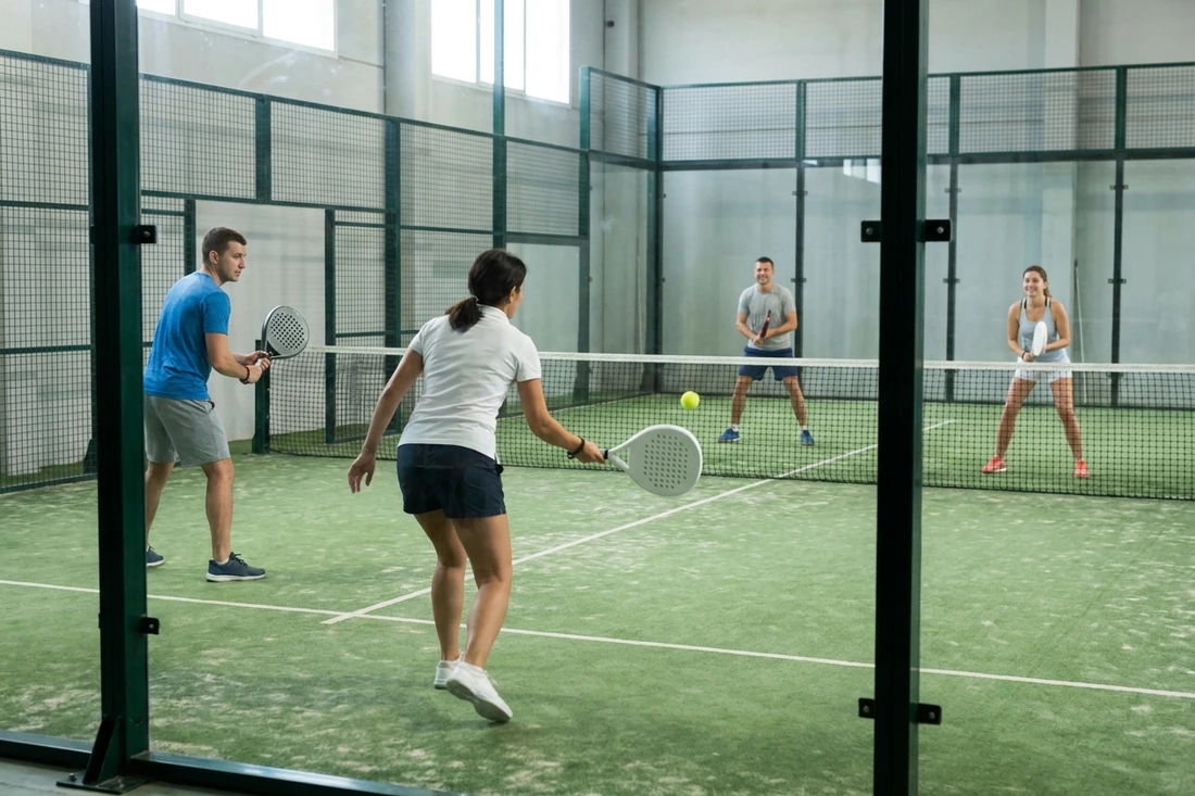 Partie de padel entre amis débutants en double, vue derrière la vitre sur un court indoor vert